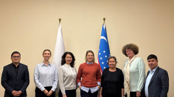 Seven individuals standing in a row in front of two flags, one of which is the flag of Uzbekistan