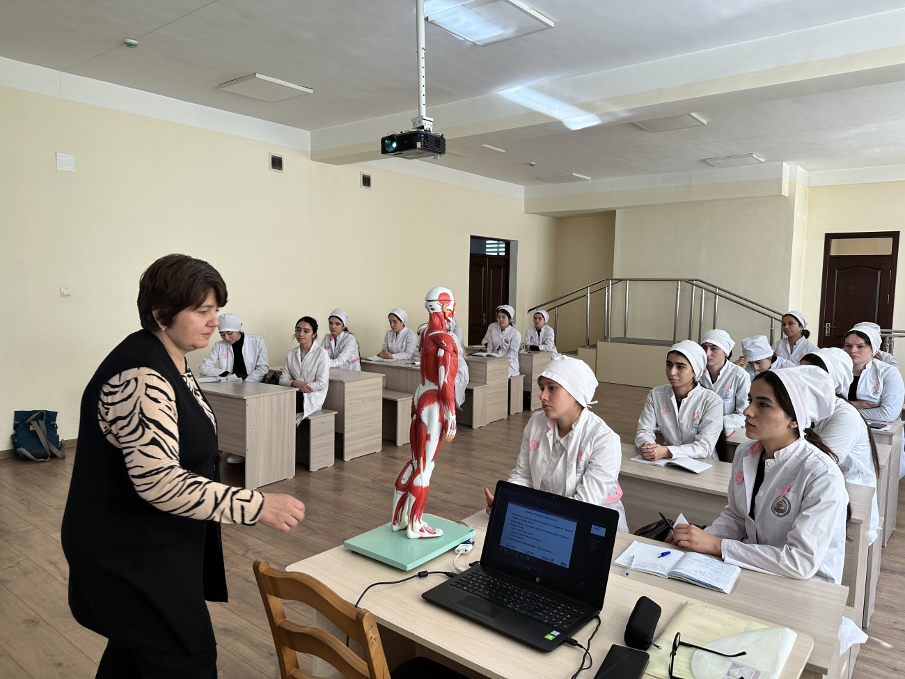 A physiotherapy instructor stands at the front of a classroom demonstrating a human muscle anatomy model while students in white medical uniforms sit at desks listening and taking notes.