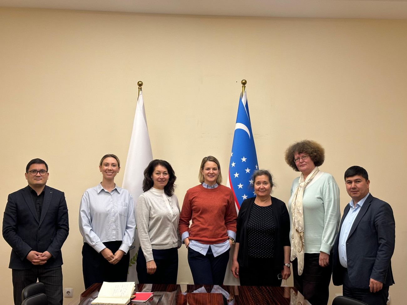 Seven individuals standing in a row in front of two flags, one of which is the flag of Uzbekistan