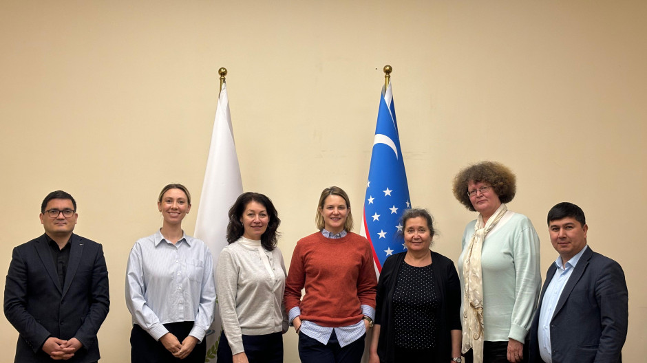 Seven individuals standing in a row in front of two flags, one of which is the flag of Uzbekistan